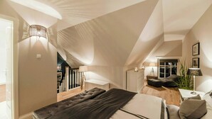 Ein Schlafzimmer mit schrägen Wänden, einem Bett, Nachttischen, einer Lampe, einer Pflanze und Blick auf eine Treppe.