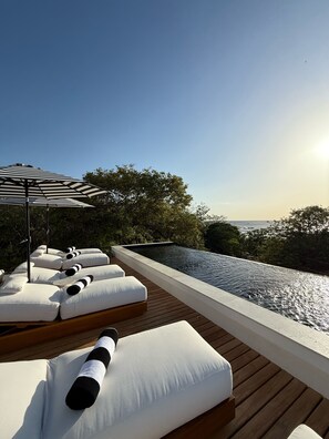 Ein Infinity-Pool auf dem Dach mit weißen Liegestühlen und einem gestreiften Sonnenschirm.
