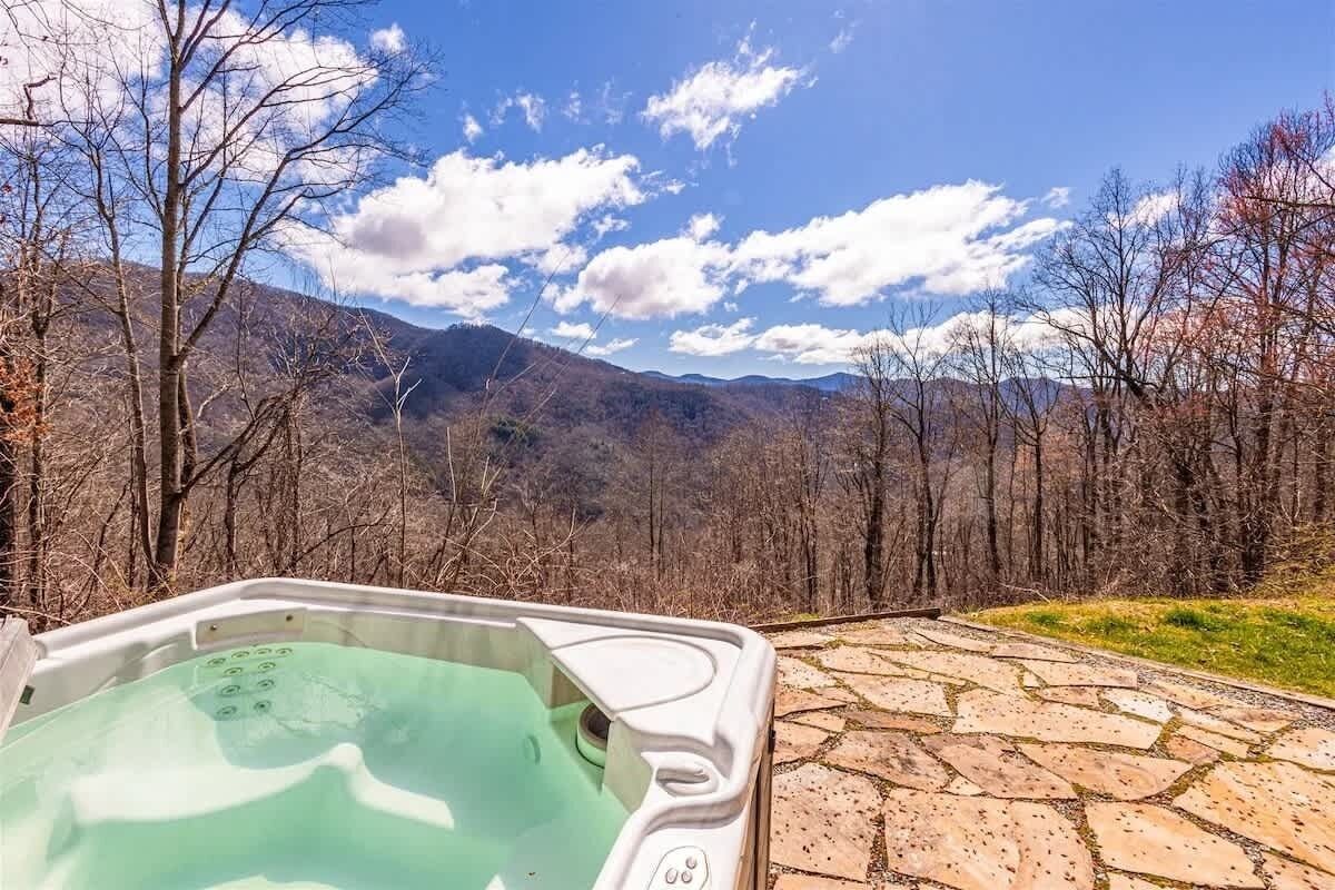 Una vasca idromassaggio con un patio in pietra, circondata da alberi e montagne.