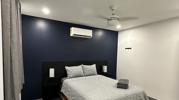 Une chambre moderne avec un mur d’accent bleu foncé, un lit blanc avec une couvre-lit à motifs, un ventilateur de plafond et un appareil de climatisation.