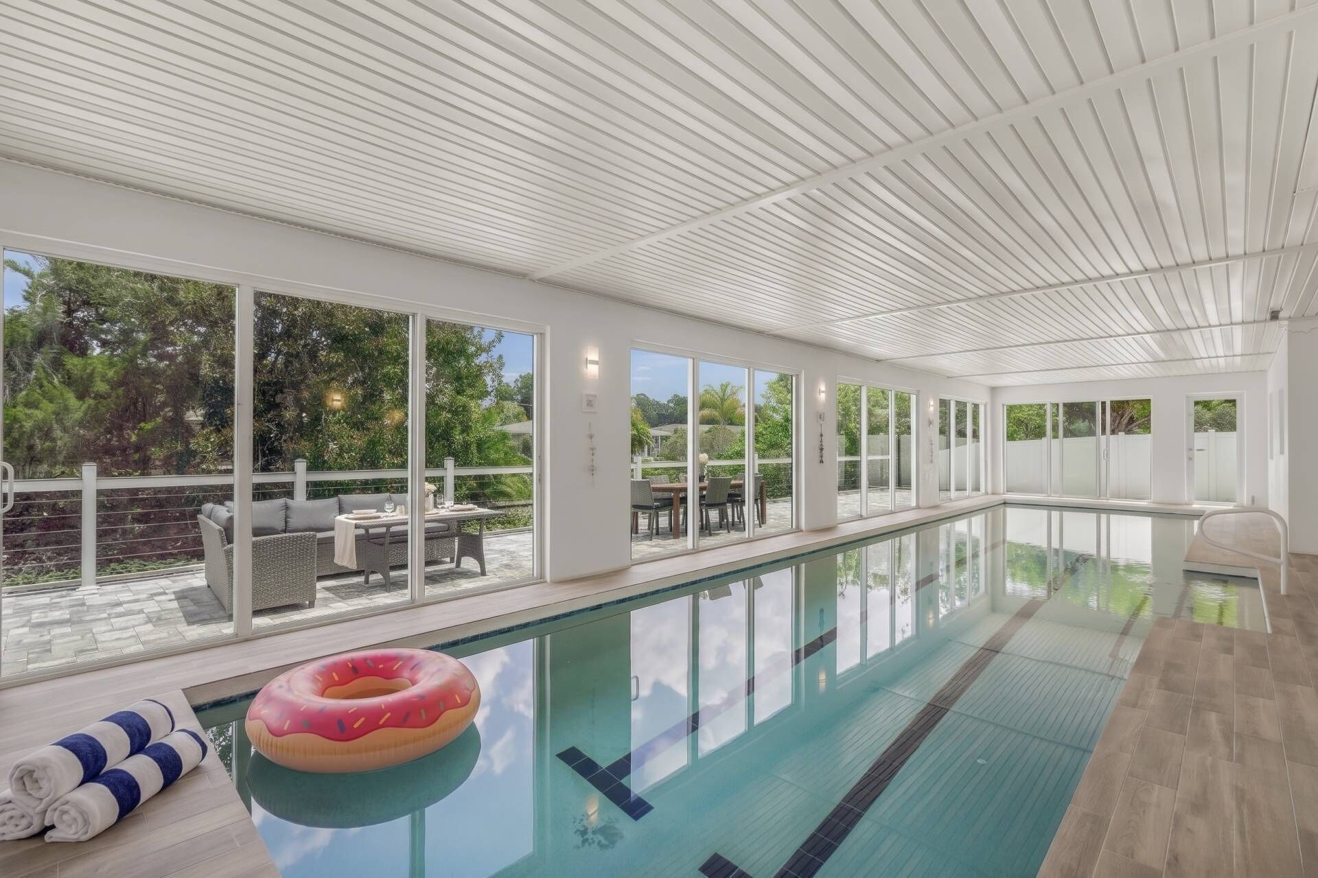 An indoor swimming pool with clear glass walls, a poolside seating area, and a view of the outdoors.