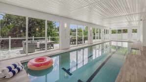 An indoor swimming pool with clear glass walls, a poolside seating area, and a view of the outdoors. - Siesta Swim House (Sarasota)