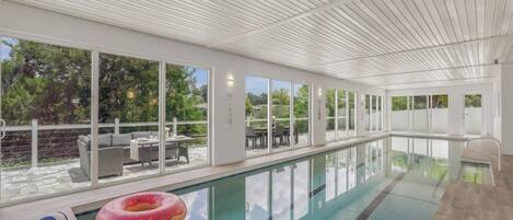 An indoor swimming pool with clear glass walls, a poolside seating area, and a view of the outdoors.