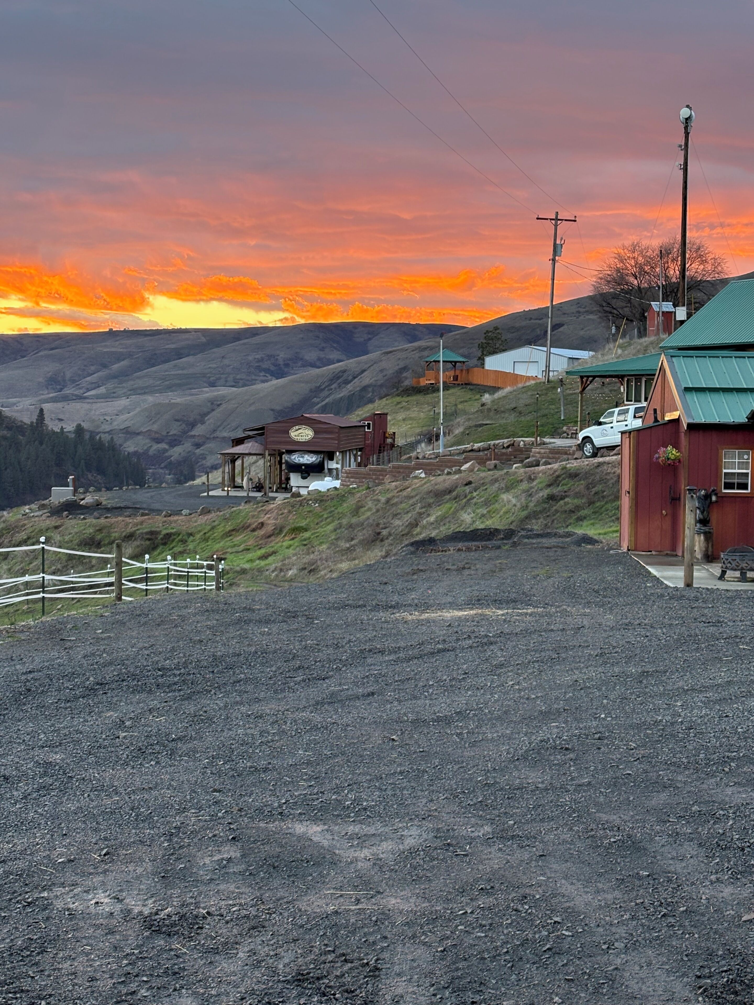 A rural landscape with a gravel road, a wooden fence, and a red building with a green roof.