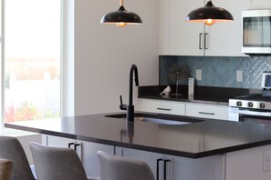 Une cuisine moderne dotée d’un îlot central, de luminaires suspendus noirs et offrant une vue sur l’extérieur.