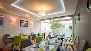 Un restaurant aux murs lambrissés, avec des chaises vertes et grises, des tables ornées d’assiettes et de tasses blanches, et une vue sur l’extérieur.