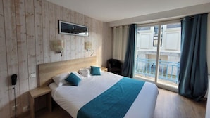 Une chambre d’hôtel avec un mur en bois, un lit avec une literie blanche et turquoise, une table de chevet avec une lampe, et une vue sur un balcon.