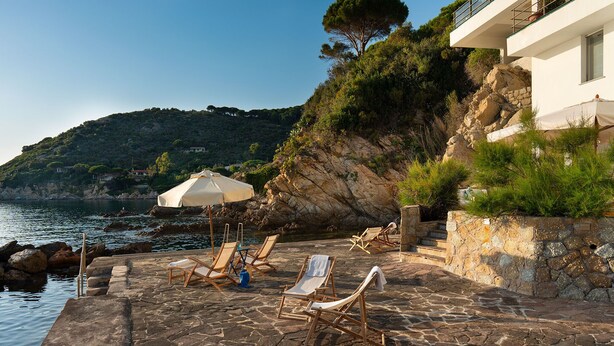 A stone patio with lounge chairs and an umbrella, overlooking a body of water and a hillside. - Villa Il Faro 10, Emma Villas (Scaglieri)