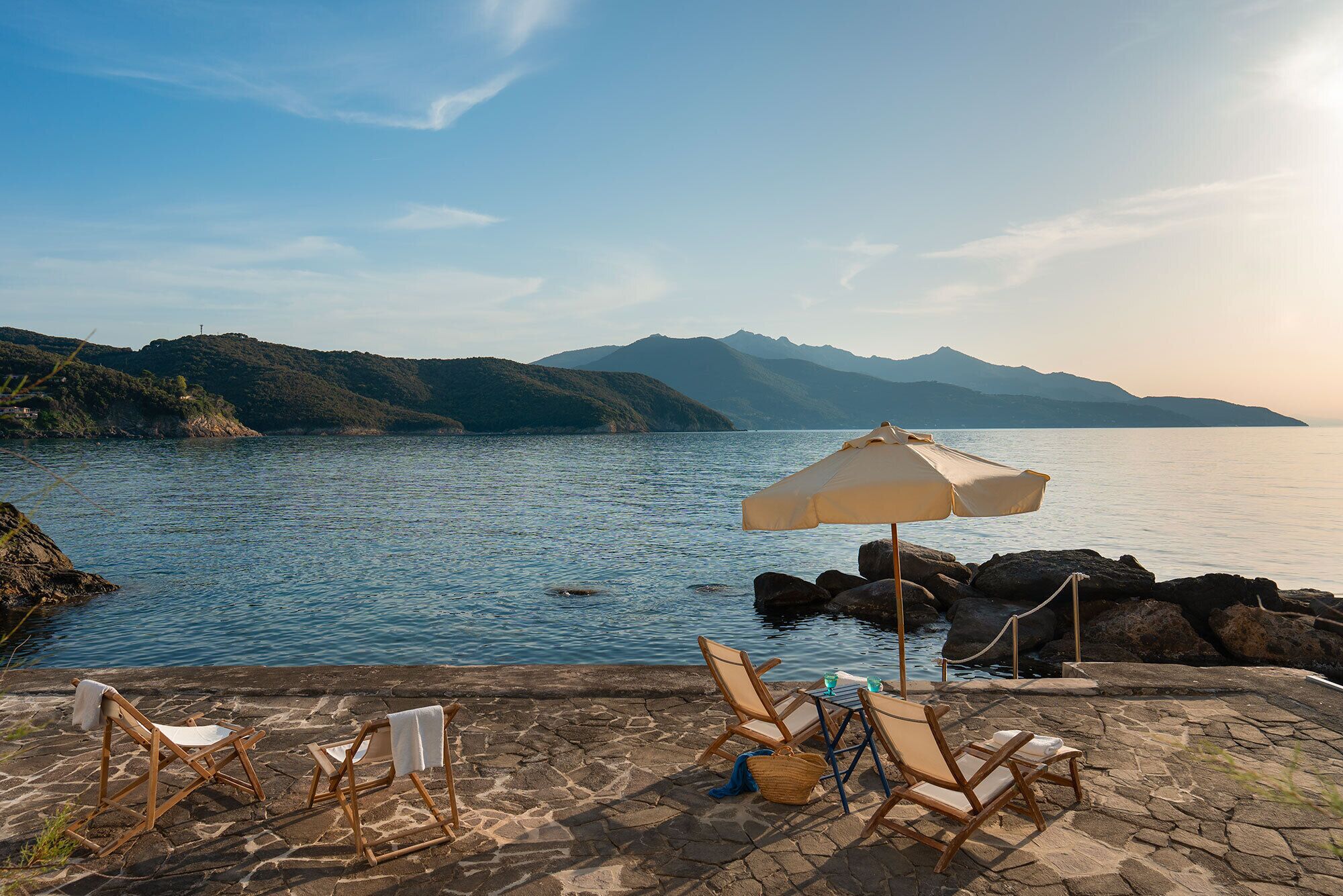 A stone patio with lounge chairs and an umbrella overlooking a calm lake with mountains in the background.