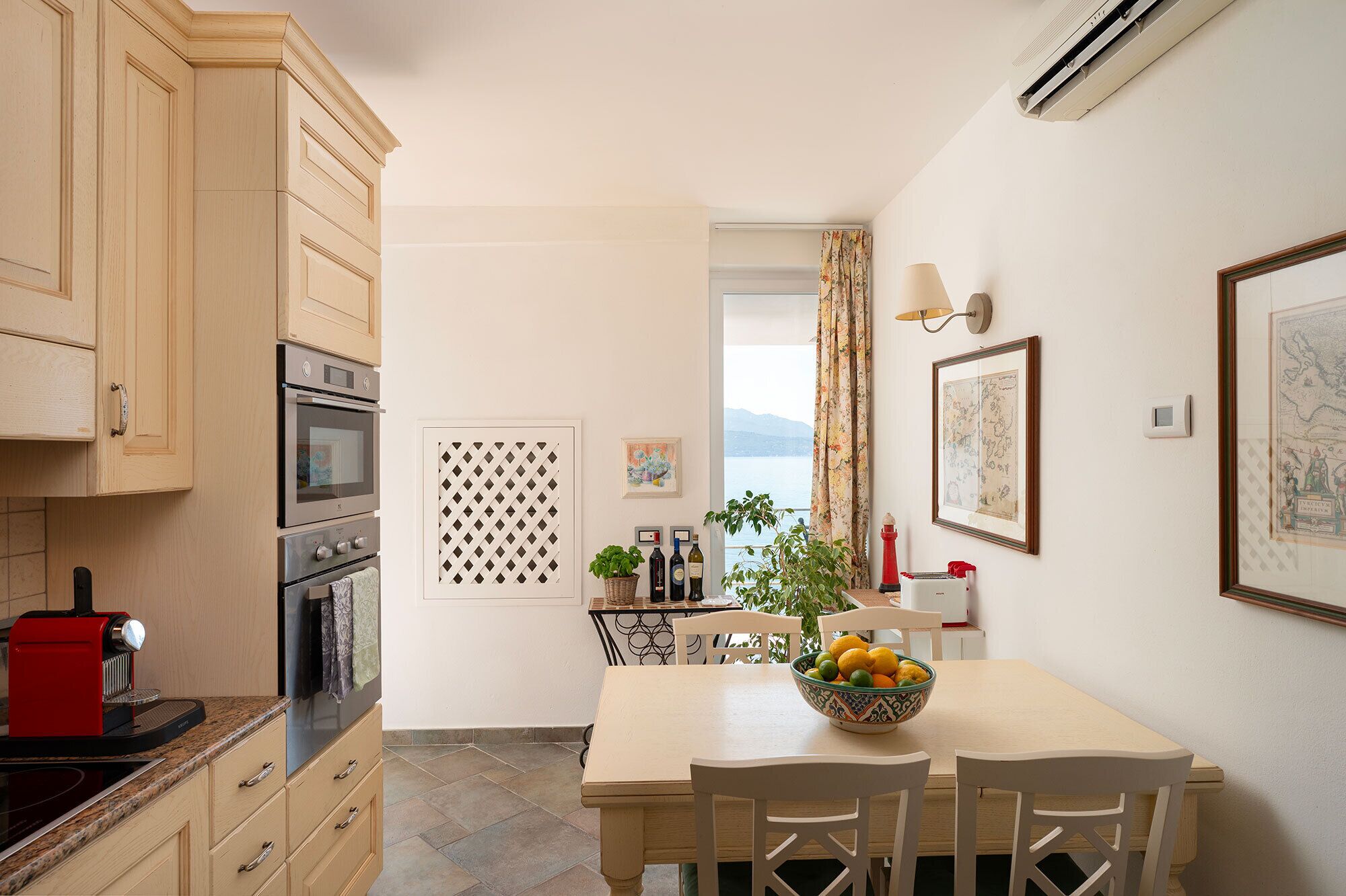A kitchen with wooden cabinets, a stove, and a red coffee machine. There is a dining table with chairs and a bowl of fruit.