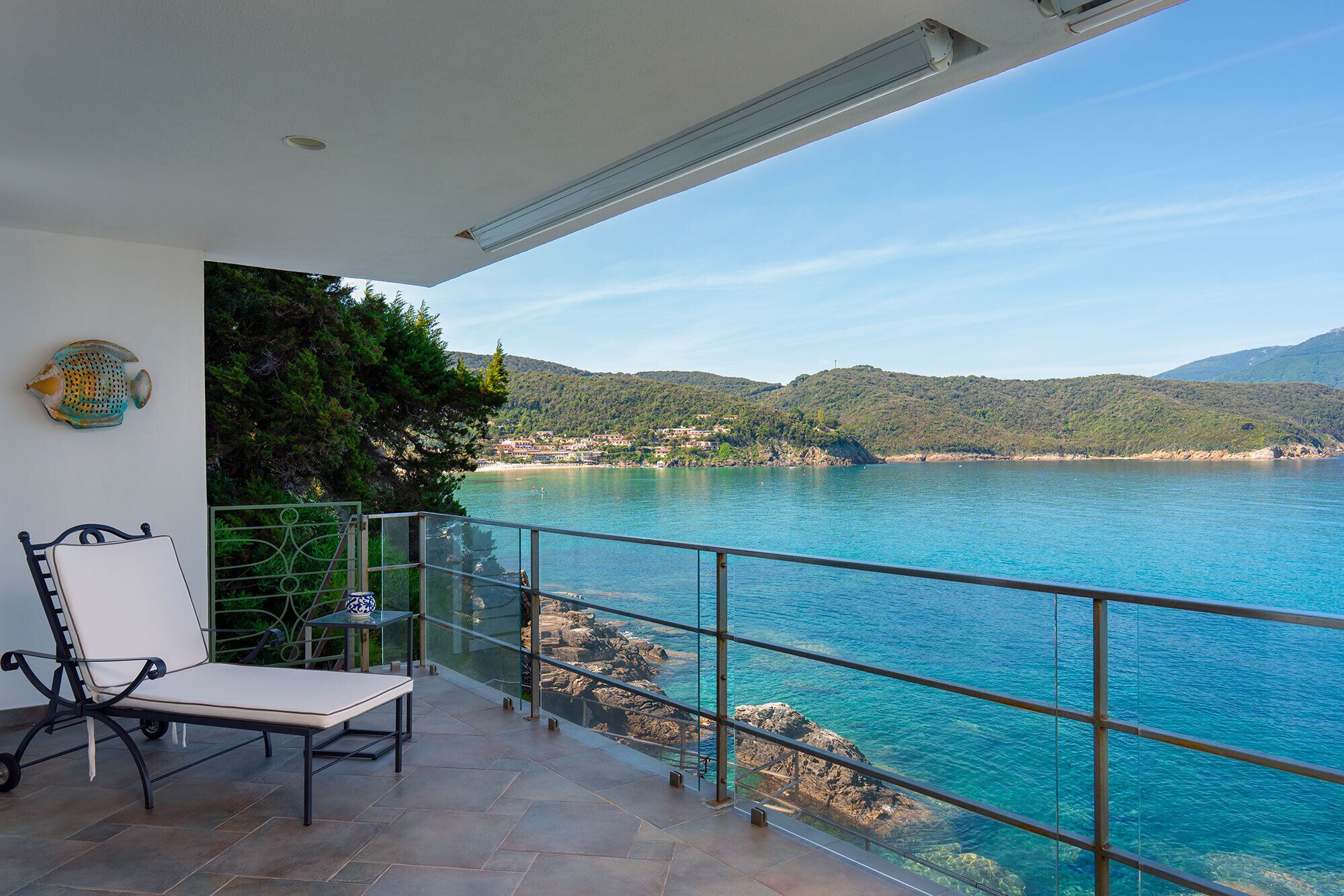 A balcony with a chair and table, overlooking a coastal area with clear blue water and hills in the distance.