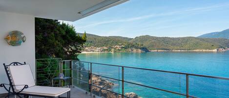 A balcony with a chair and table, overlooking a coastal area with clear blue water and hills in the distance.