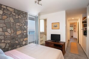 A bedroom with a stone wall, a bed, a television on a wooden dresser, and a hallway leading to a bathroom. - Villa Il Faro 10, Emma Villas (Scaglieri)