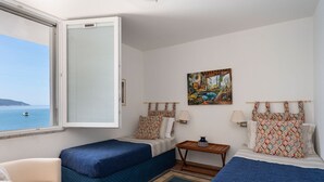 A hotel room with two beds, a sofa, a small table, a painting, and a view of the sea. - Villa Il Faro 10, Emma Villas (Scaglieri)