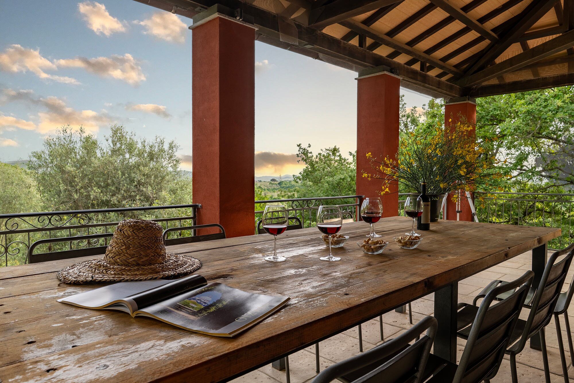 A wooden table with wine glasses and a bottle, a woven basket, and a magazine on a terrace with red pillars and a view of greenery.