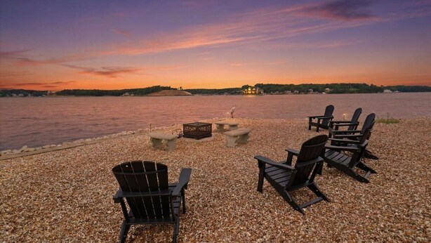 A pebbled beach with Adirondack chairs and a fire pit, overlooking a body of water at sunset. - 19MM w/ Dock, Boat & PWC Slips, Views, Game Room, Fire Pit & 2-Story Deck (Osage Beach)