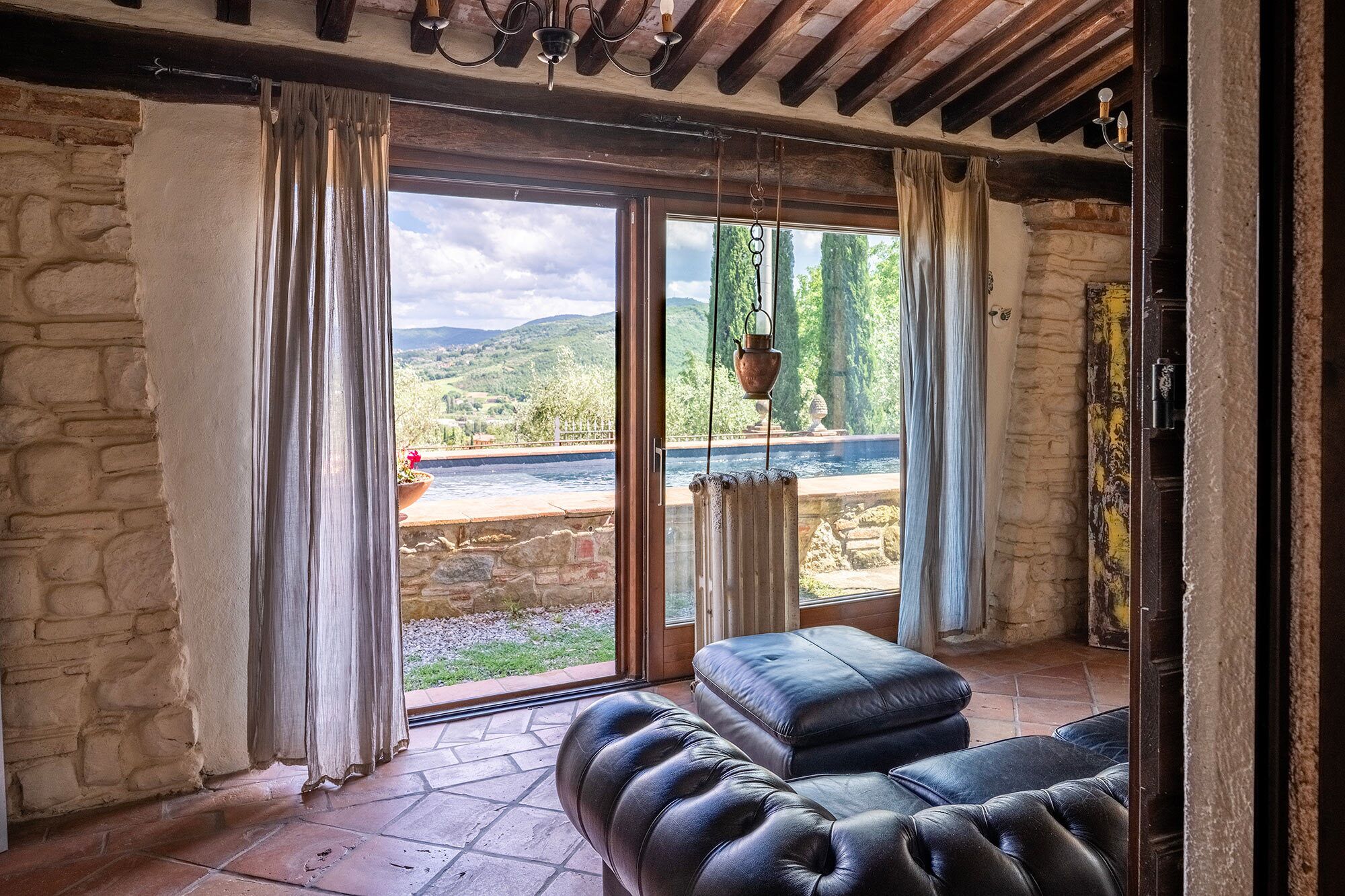 Ein Zimmer mit einer Steinwand, einem großen Fenster mit Vorhängen, einem schwarzen Ledersofa und einer hängenden Pflanze.