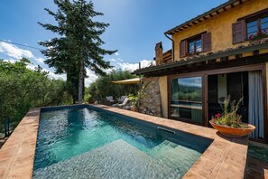 A swimming pool with a stone border, a stone wall, and a wooden deck with outdoor furniture. - Colle Calzolaro 8, Emma Villas (Tavernelle)