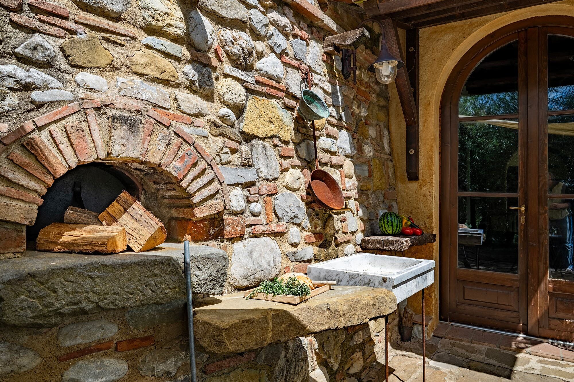 A stone building with a brick oven, a wooden shelf with vegetables, and a stone bench.