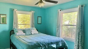 Quarto com cama, duas janelas, ventilador de teto e vista para árvores.