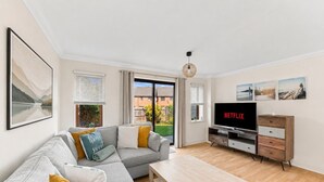 Una sala de estar moderna con un sofá gris, una mesa de centro de madera, un televisor mostrando 'NETFLIX' y vistas a un jardín a través de puertas correderas.