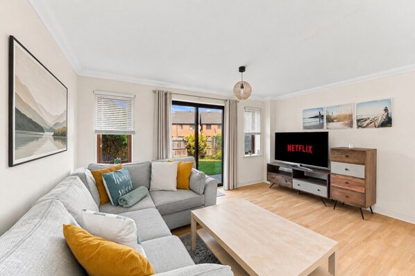 Una sala de estar moderna con un sofá gris, una mesa de centro de madera, un televisor mostrando 'NETFLIX' y vistas a un jardín a través de puertas correderas.