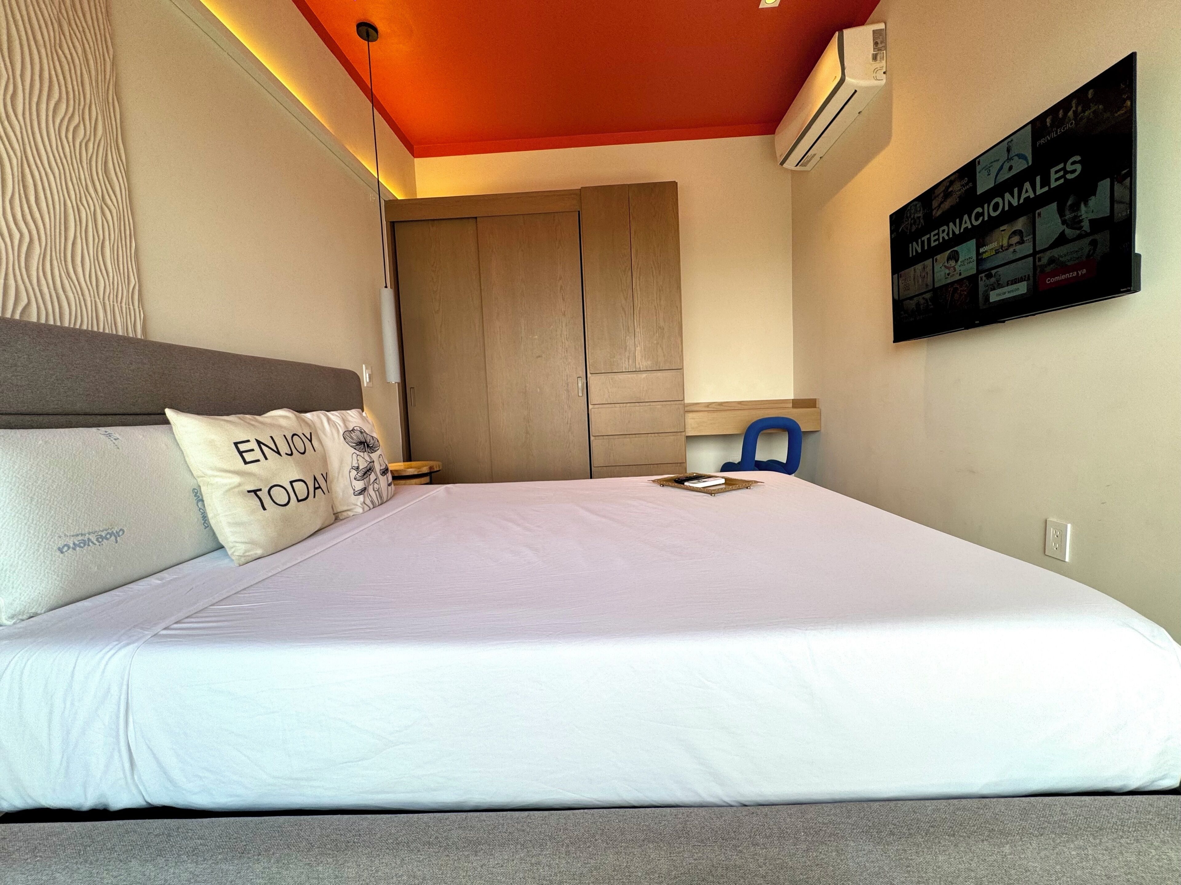 A bedroom with a bed, pillows, a nightstand, a chair, a wardrobe, and a wall-mounted TV displaying a program.