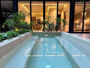 Una piscina interior rectangular con paredes y suelo de baldosas, rodeada de plantas en macetas y una zona de estar con sillones.