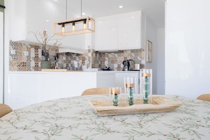 Una cocina moderna con armarios blancos, salpicadero estampado y una zona de comedor con mantel floral.