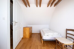 Ein kleines Zimmer mit einem Einzelbett, einem Holzkommode, einem Stuhl und einer Tür zu einem weiteren Raum.