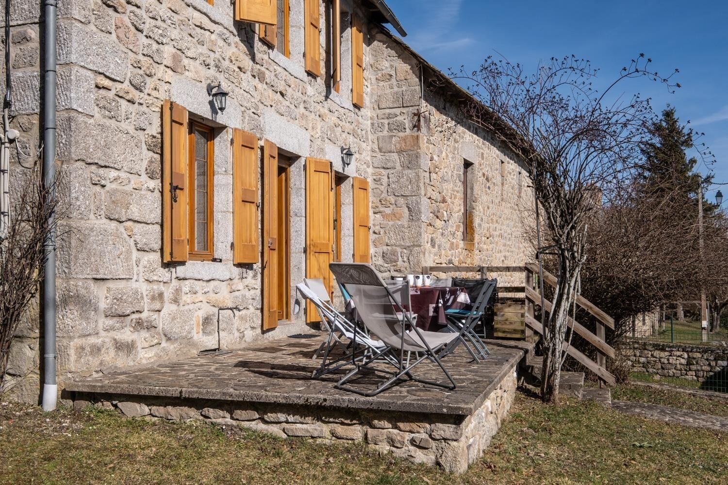 Un bâtiment en pierre avec des volets en bois, une terrasse avec une table et des chaises, et un escalier menant à un niveau supérieur.
