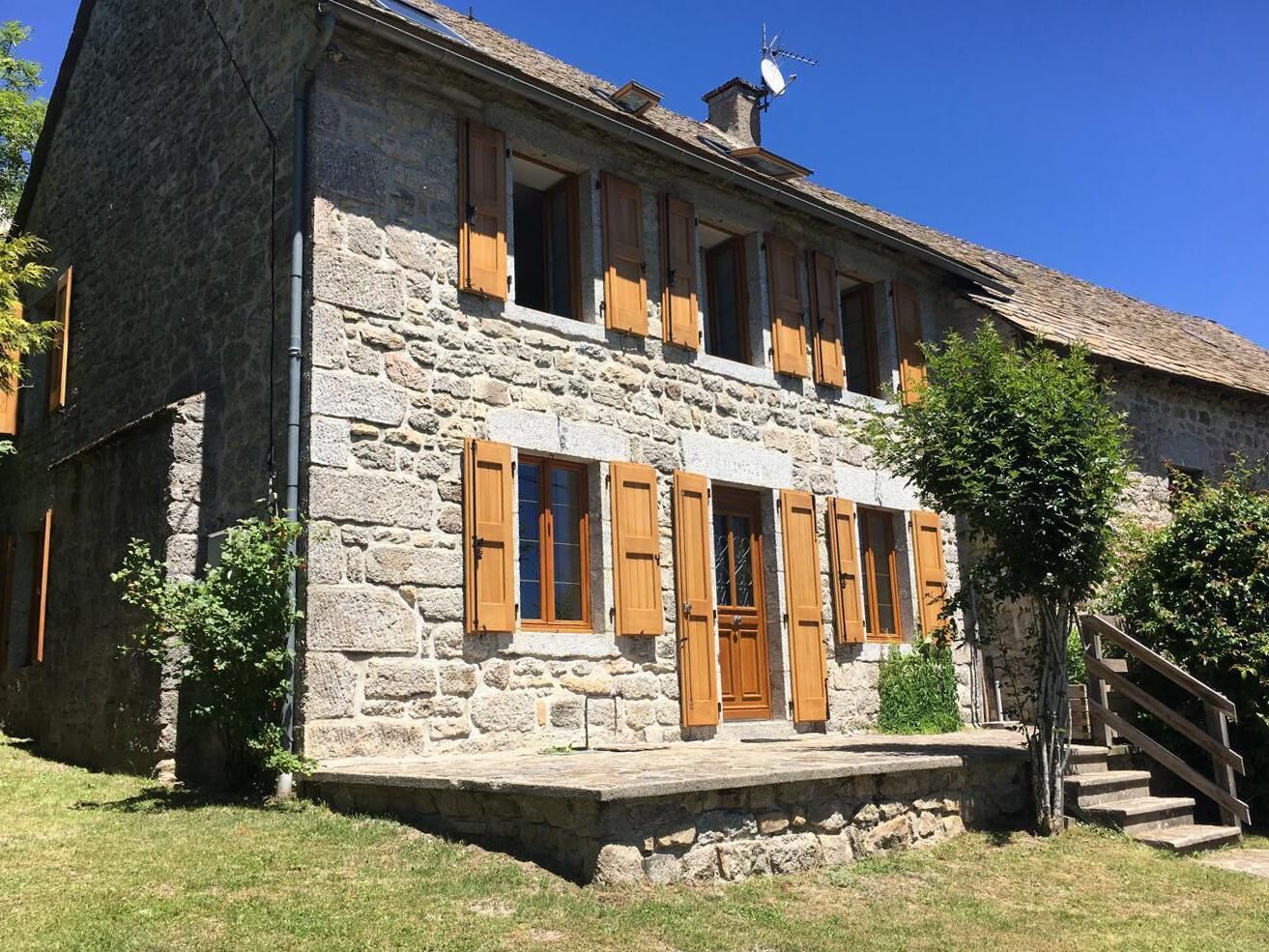 Un bâtiment en pierre avec des volets en bois, une cheminée et un escalier menant à l’entrée.