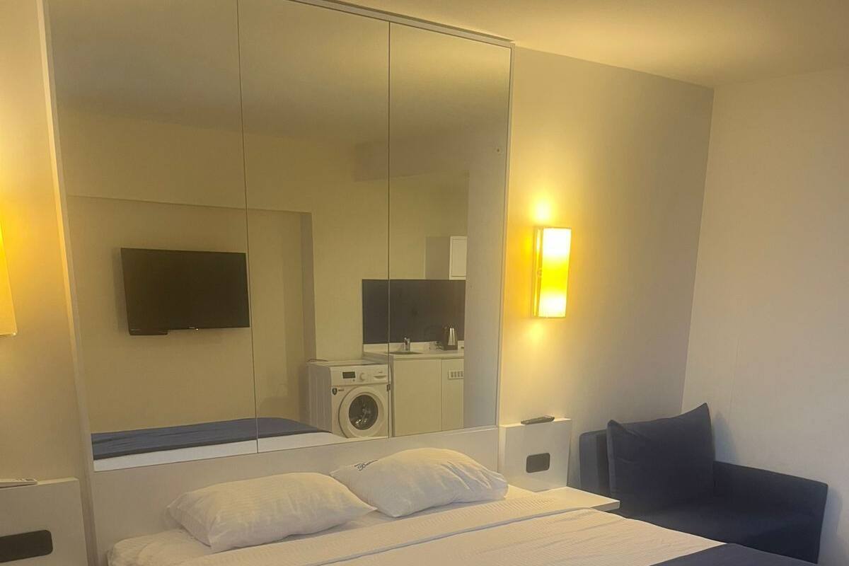 A modern hotel room with a large bed, a built-in washing machine, a flat-screen TV, and a bedside table.
