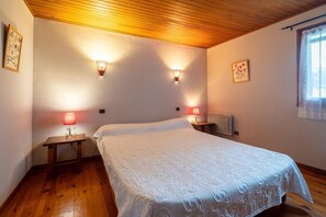 Ein Schlafzimmer mit Holzbalkendecke, zwei Nachttischlampen, einem Bett mit weißer Bettdecke und einem Fenster mit Vorhang.