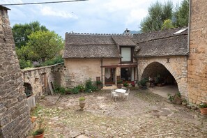 A stone building with a tiled roof, a courtyard with a table and chairs, and an archway leading to a covered area. - Domaine Rivière (6–7 people) (BRENOUX)