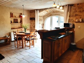A cozy kitchen with wooden cabinets, a dining table with chairs, a hanging light fixture, and a window with curtains. - The Maison Caussenarde at La Bazalgette (SAINT ETIENNE DU VALDONNEZ)