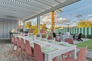 En udendørs spiseplads med en overdækket terrasse, grill og et bord dækket til seks personer med pink stole.