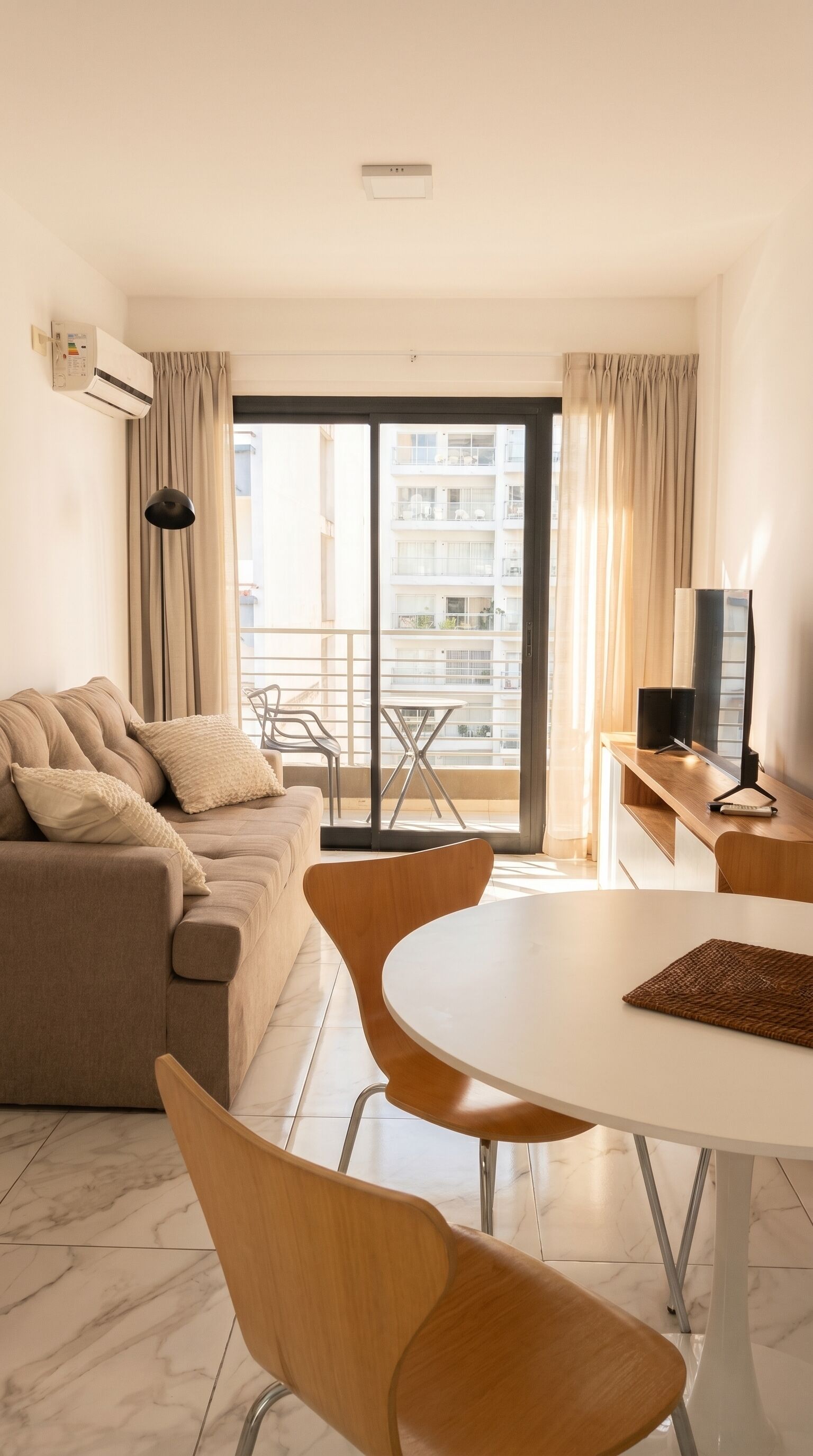 Ein modernes Wohnzimmer mit einer Couch, einem runden Tisch und Stühlen. Ein Balkon bietet Blick auf Gebäude.