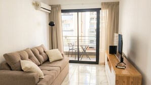 A living room with a sofa, a TV on a wooden stand, a balcony with a table and chairs, and an air conditioning unit on the wall.