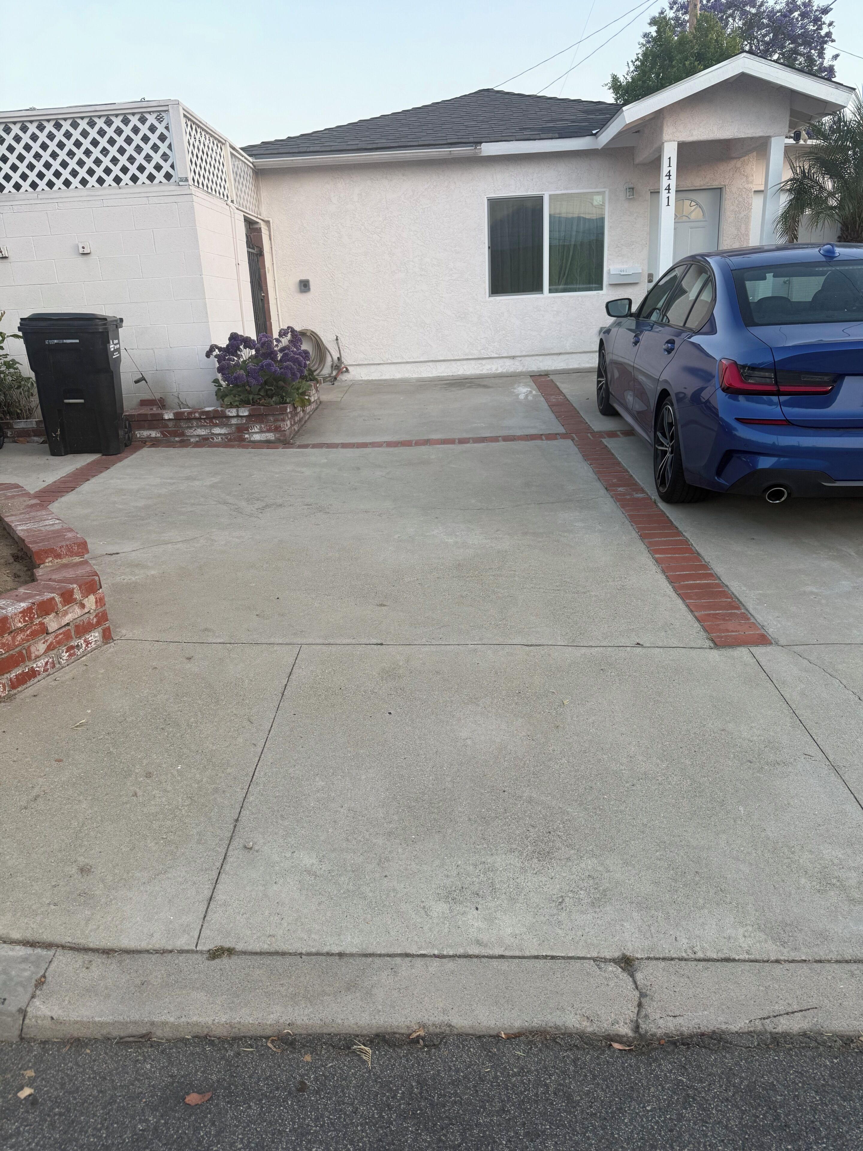 Un camino de entrada residencial con un coche azul estacionado a la derecha, una casa blanca con el número '1441' en la fachada, y un contenedor de basura negro a la izquierda.