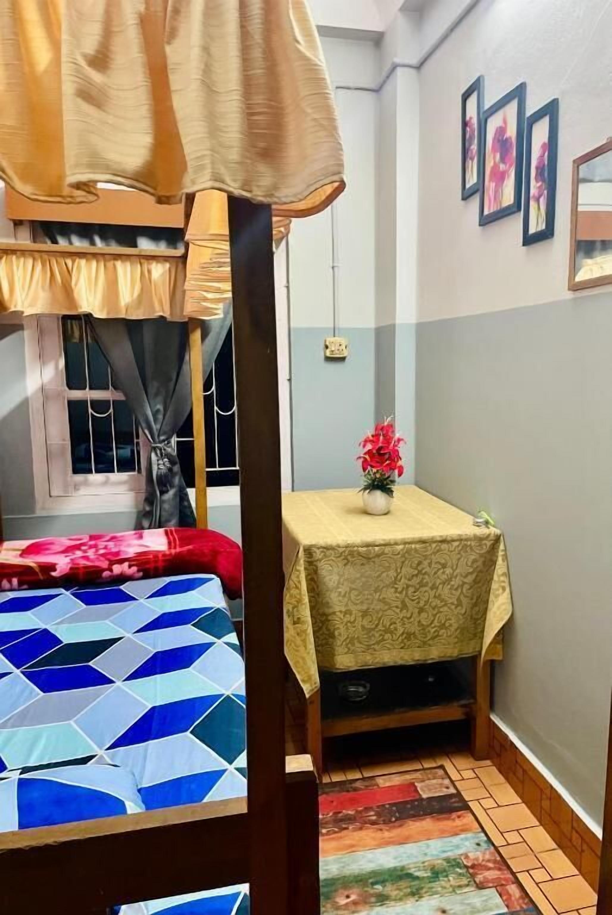 A room with a bunk bed, a small table with a tablecloth, a potted plant, and framed pictures on the wall.