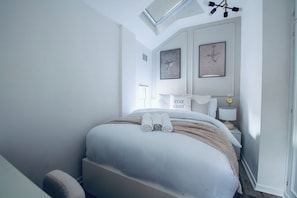 A bedroom with a bed, pillows, a bedside table, a lamp, and artwork on the wall.