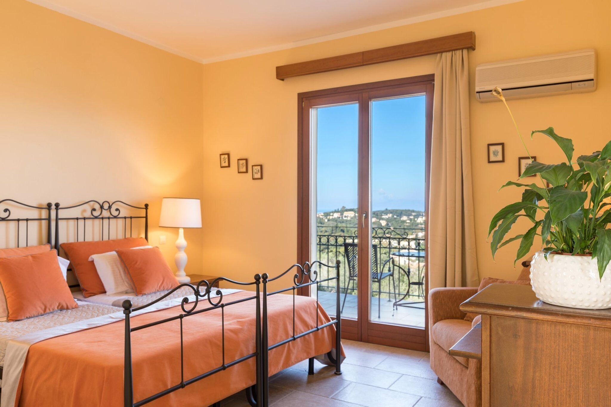 Ein Schlafzimmer mit Bett, Nachttisch, Lampe, einem Topf mit Pflanze und einem Balkon mit Aussicht.