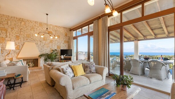 A living room with a stone wall, a sofa, a TV, and a balcony with wicker furniture. - Villa Melioris, Divine Sea Views, Private Pool, Spacious Furnished Terrace,. (Corfu)