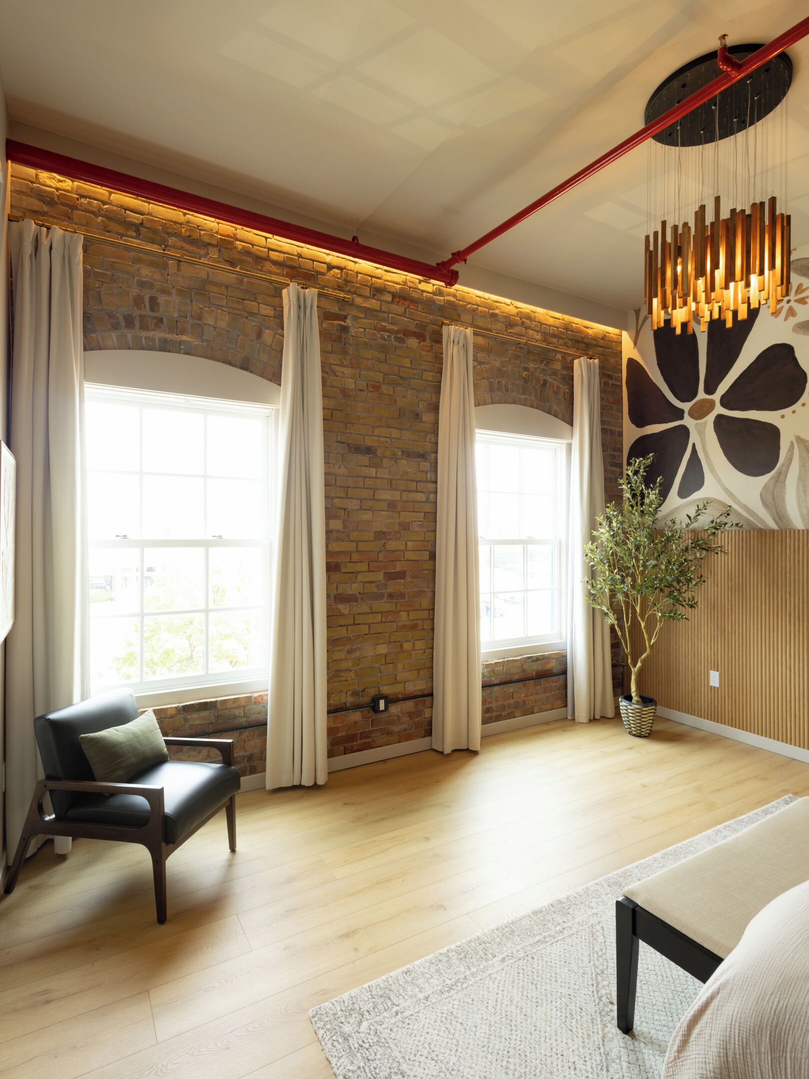 A room with a brick wall, large windows with curtains, a black leather chair, a wooden floor, and a hanging light fixture.