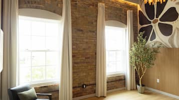 A room with a brick wall, large windows with curtains, a black leather chair, a wooden floor, and a hanging light fixture.