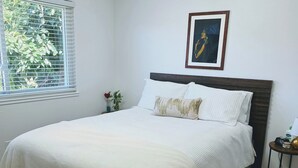 A bedroom with a bed, a window with blinds, a framed picture on the wall, and a small table with a lamp.