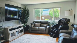 A living room with a flat-screen TV, leather sofas, a massage chair, a large window, and a bookshelf.