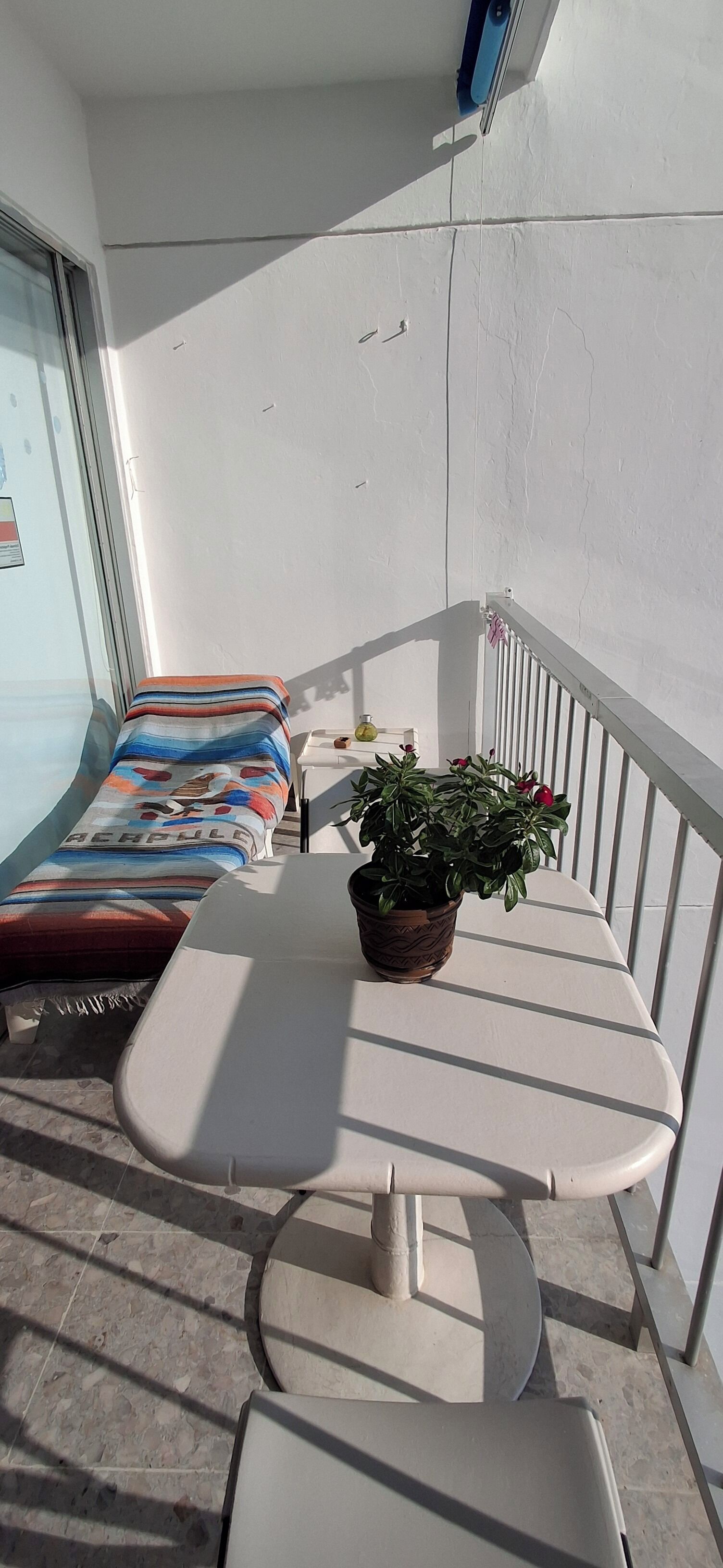 A balcony with a white table, a potted plant, and a bed with striped bedding.