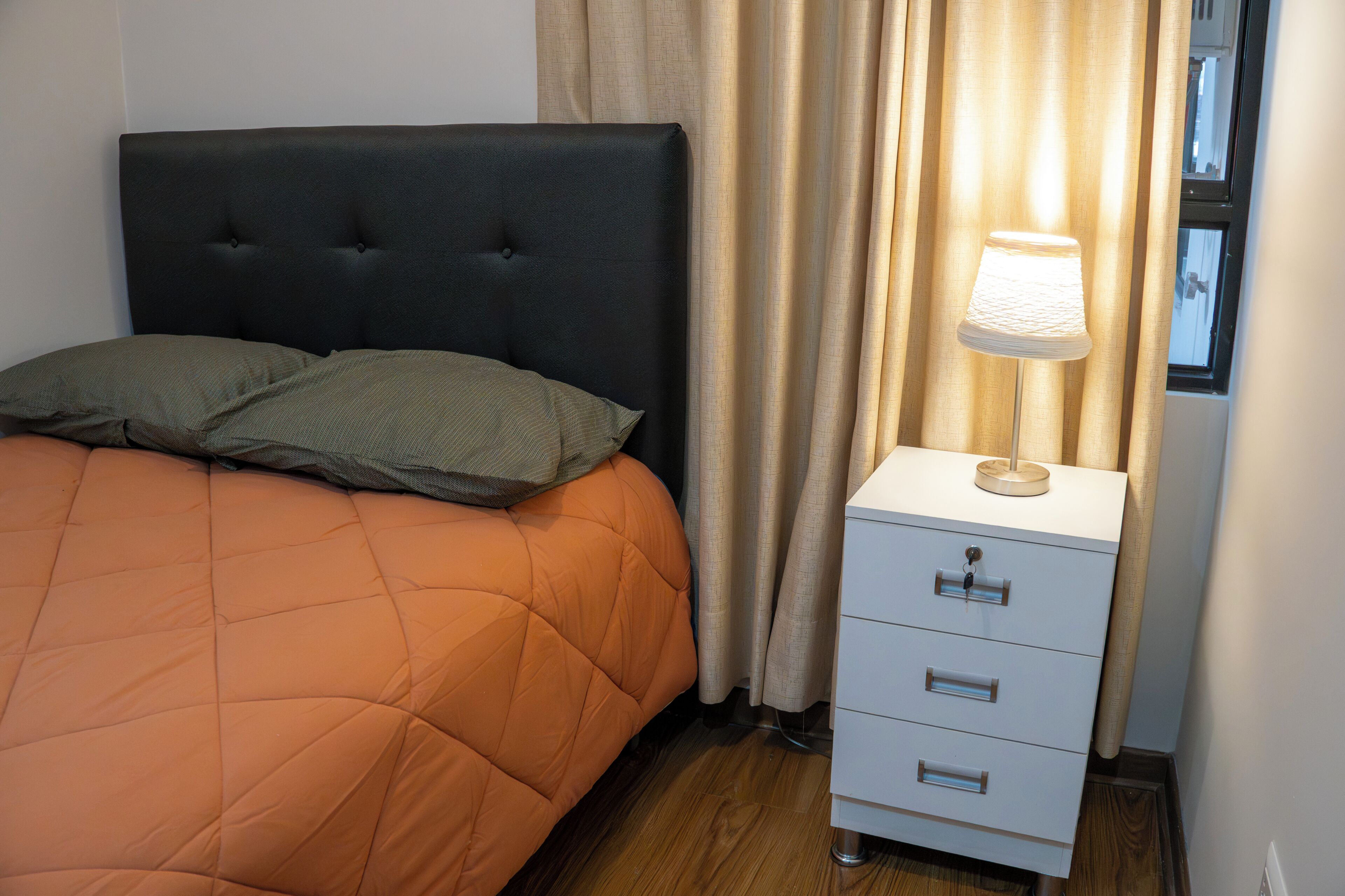 A bed with an orange quilt, a black headboard, and two pillows. There is a bedside table with a lamp and a drawer.